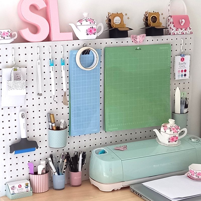 A beech wood desk with a Cricut Explore Air 2 digital cutting machine on top and a white pegboard with papercrafting accessories hanging from it