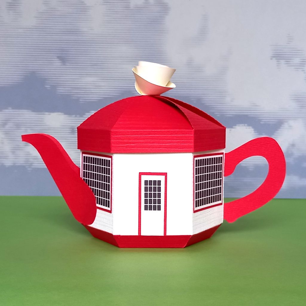 A small papercraft teapot gift box made in the style of the teapot dome gas station with a white body a red lid a red handle and spout and tiny black windows