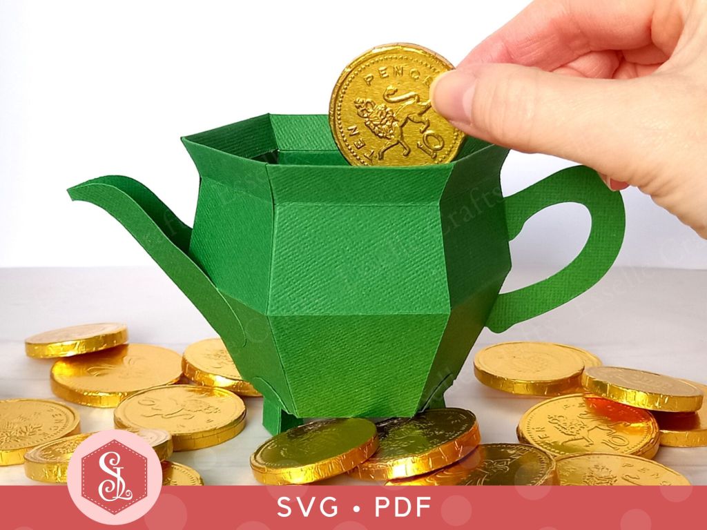 A green papercraft teapot gift box in the shape of a St Patricks Day pot of gold surrounded by gold chocolate coins. One gold coin is being placed inside the top of the box.