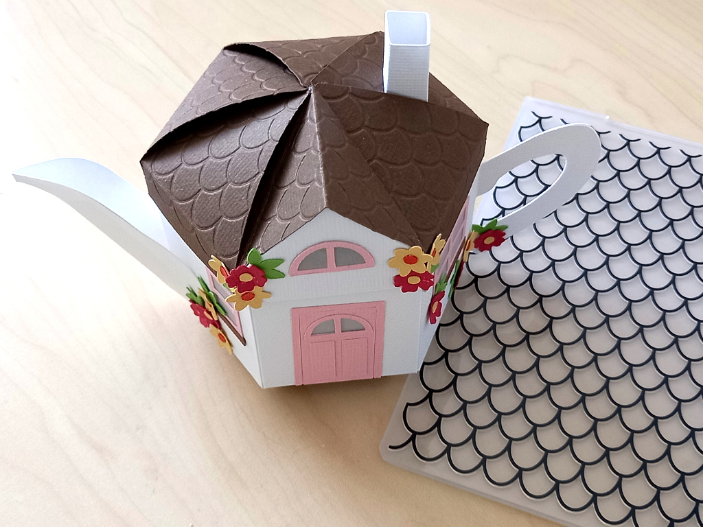 A 3D paper craft teapot gift box made in the style of a cosy country cottage with a pink door and pink windows decorated with small paper flowers