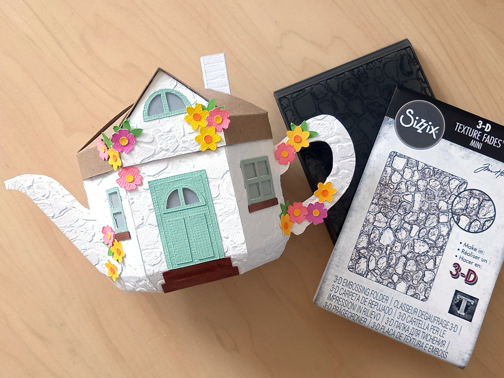 A 3D paper craft teapot gift box made in the style of a cosy country cottage with a mint green door and windows decorated with small paper flowers