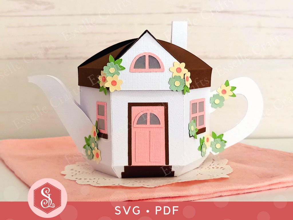 A 3D paper craft teapot gift box made in the style of a cosy country cottage with a pink door and pink windows decorated with small paper flowers