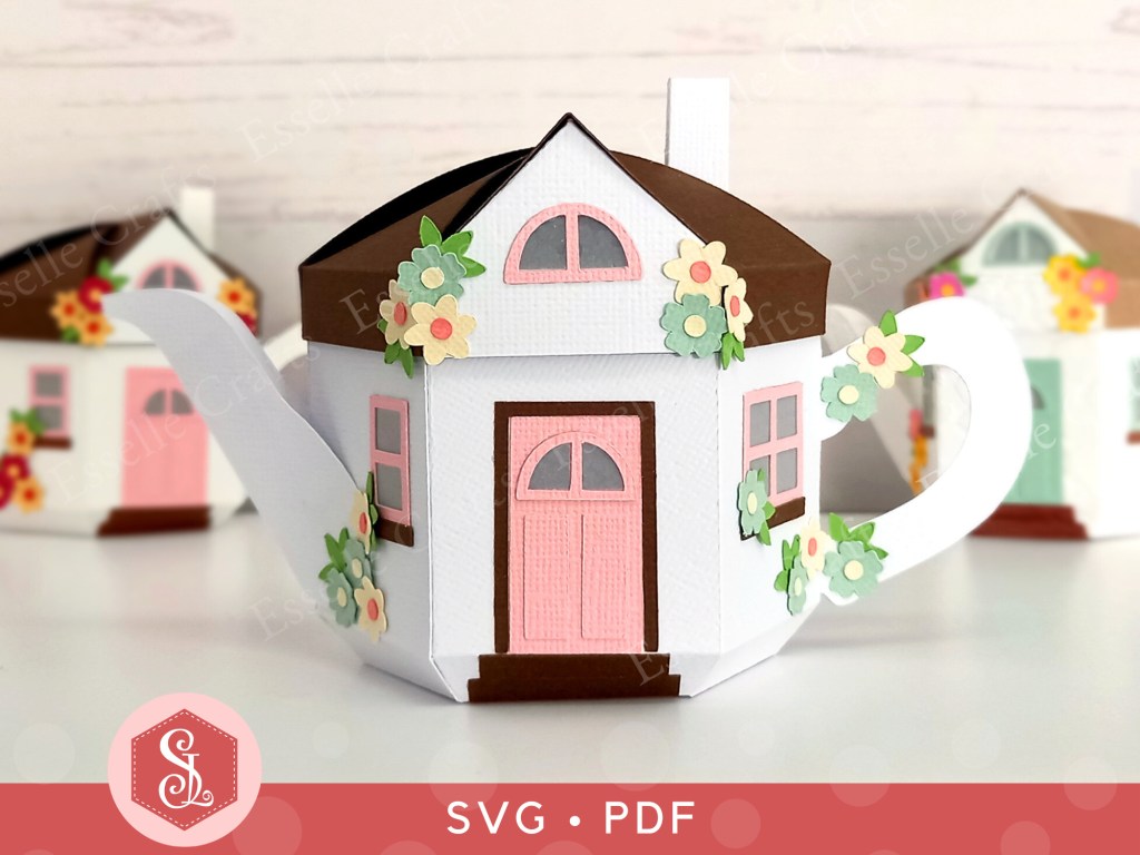 A 3D paper craft teapot gift box made in the style of a cosy country cottage with a pink door and pink windows decorated with small paper flowers