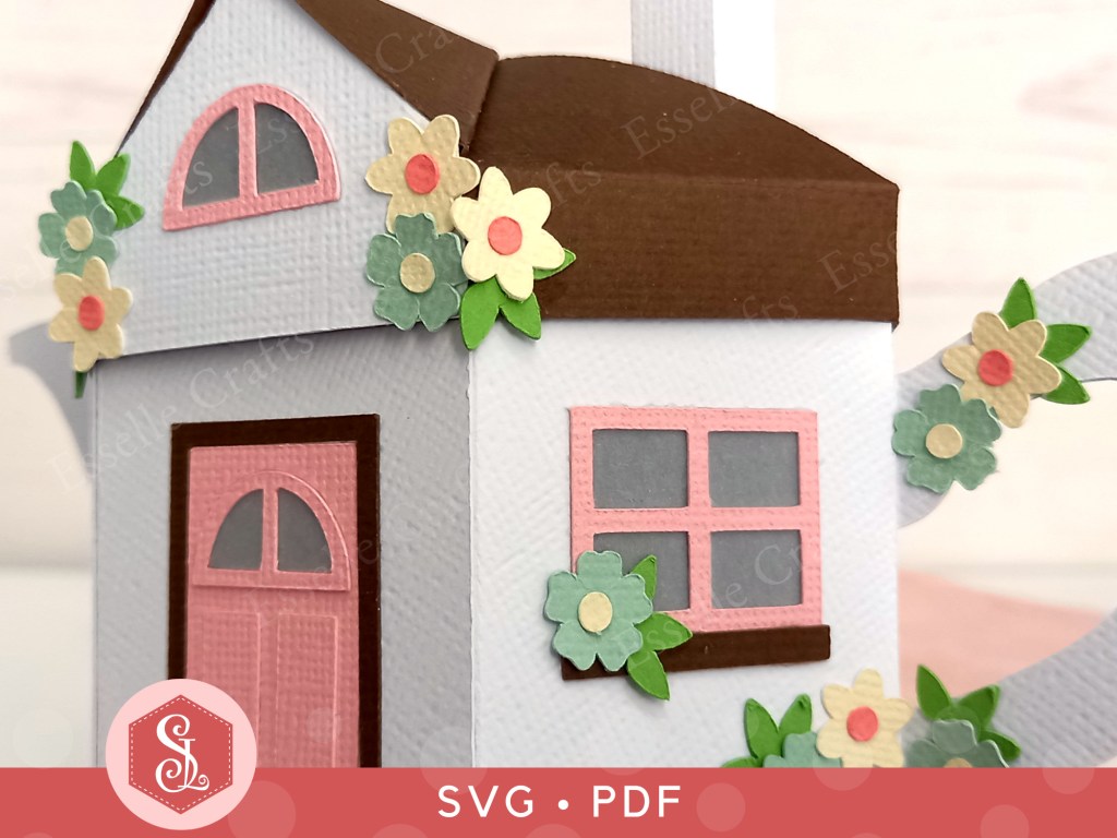 A 3D paper craft teapot gift box made in the style of a cosy country cottage with a pink door and pink windows decorated with small paper flowers