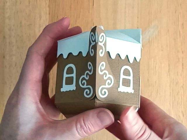 A brown paper craft teapot gift box in the style of a classic gingerbread house with a white icing lid white icing windows and white icing swirls