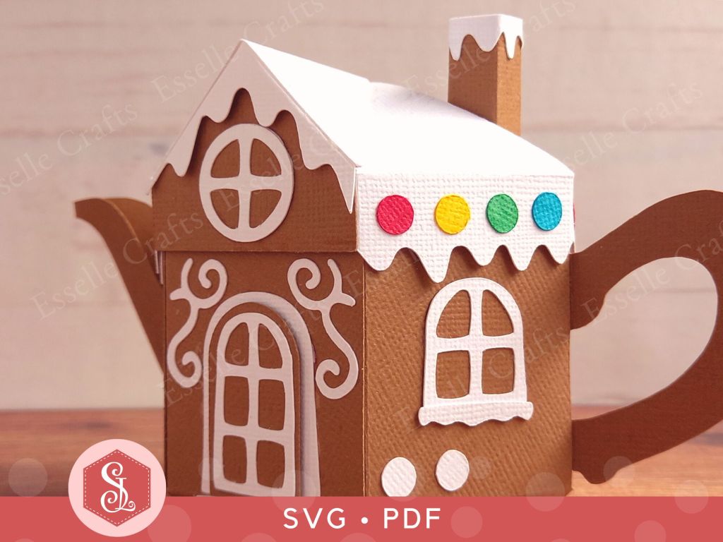 A brown paper craft teapot gift box in the style of a classic gingerbread house with a white icing lid white icing door and windows and white icing swirls