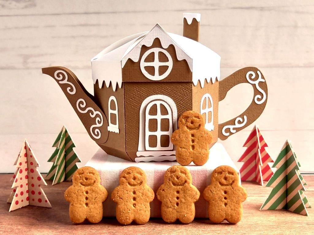A brown paper craft teapot gift box in the style of a classic gingerbread house with white icing door and windows and white icing swirls with some mini gingerbread men around it