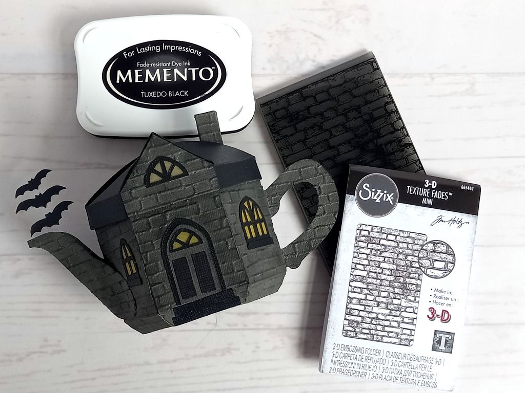 A paper craft haunted house gift box for Halloween trick or treating in the shape of a teapot with yellow arched gothic windows and three bats flying out of the spout