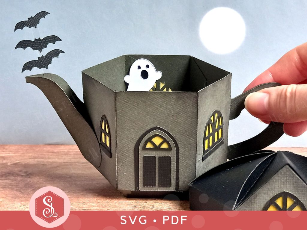 A paper craft haunted house gift box for Halloween trick or treating in the shape of a teapot with yellow arched gothic windows a white paper ghost inside and three bats flying out of the spout
