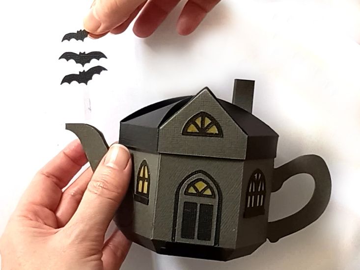 A paper craft haunted house gift box for Halloween trick or treating in the shape of a teapot with yellow arched gothic windows and three bats flying out of the spout