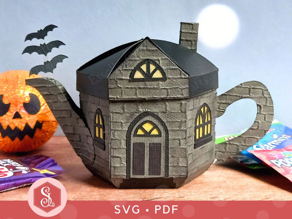 A paper craft haunted house gift box for Halloween trick or treating in the shape of a teapot with yellow arched gothic windows and three bats flying out of the spout