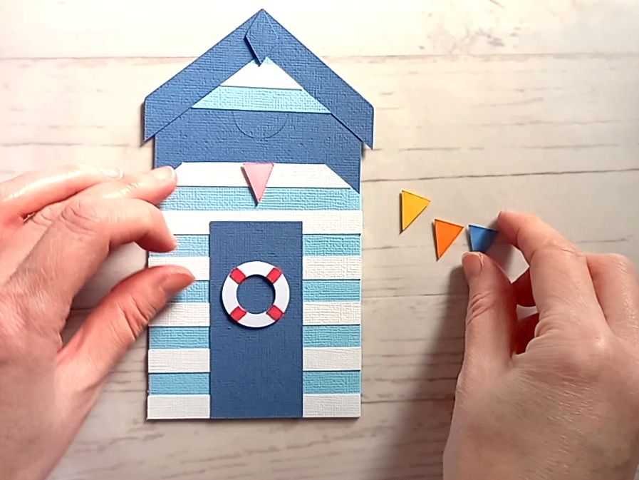 A papercraft gift card holder in the style of a beach hut, decorated with blue and white stripes, a red and white lifebuoy and colourful paper bunting.