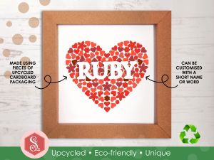 A red mosaic heart design made from small pieces of upcycled paper packaging, framed in a square brown kraft card frame. The word RUBY is displayed across the middle in white capital letters. 
