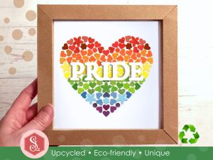 A rainbow mosaic heart design made from small pieces of upcycled paper packaging, framed in a square brown kraft card frame. The word PRIDE is displayed across the middle in white capital letters. 