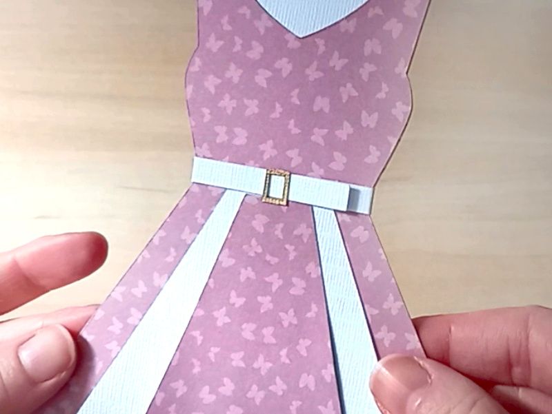 A papercraft gift card holder in the style of a vintage pink dress. It is decorated with a gold paper buckle at the waist and white paper panels on the front.