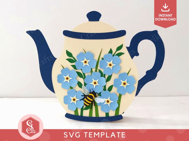 A papercraft gift card holder in the style of a vintage cream teapot with a dark blue handle and spout. It is decorated with light blue paper forget-me-not flowers on the front.