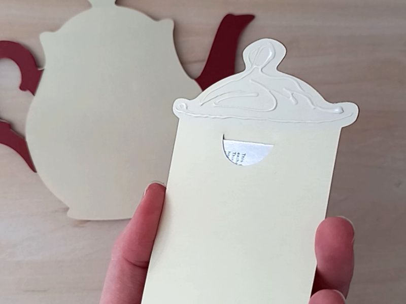 A papercraft gift card holder in the style of a vintage cream teapot with a dark red handle and spout. It is decorated with red and green paper poinsettias on the front.