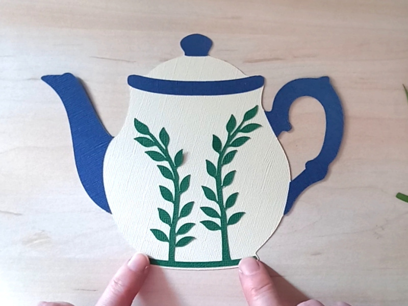 A papercraft gift card holder in the style of a vintage cream teapot with a dark blue handle and spout. It is decorated with light blue paper forget-me-not flowers on the front.