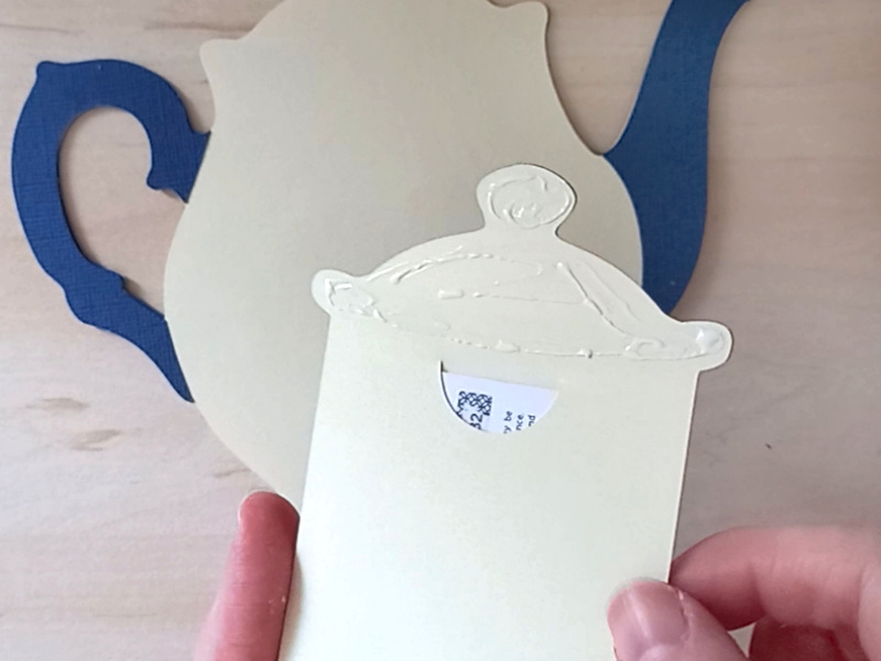 A papercraft gift card holder in the style of a vintage cream teapot with a dark blue handle and spout.