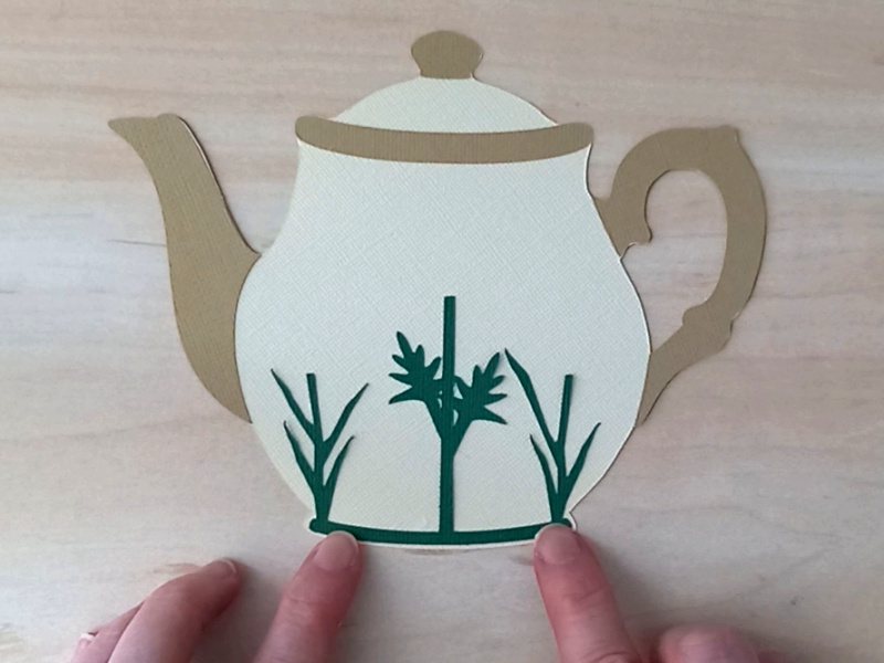 A papercraft gift card holder in the style of a vintage cream teapot with a pale brown handle and spout. It is decorated with a paper poppy, two paper cornflowers, three paper daisies and a paper bee on the front.