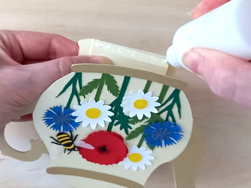 A papercraft gift card holder in the style of a vintage cream teapot with a pale brown handle and spout. It is decorated with a paper poppy, two paper cornflowers, three paper daisies and a paper bee on the front.