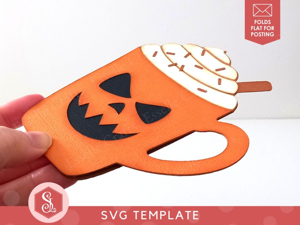 A papercraft gift card holder in the style of a pumpkin spice latte mug. It has a spooky black cut out face in the centre of the mug and is topped with white paper whipped cream, a brown paper cinnamon stick and dark brown paper sprinkles.