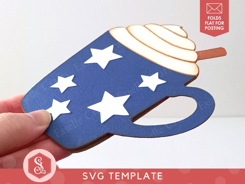 A papercraft gift card holder in the shape of a cosy Christmas mug. It is dark blue, decorated with white paper stars, and is topped with white paper whipped cream and a brown paper cinnamon stick. It is folded flat for easy posting.